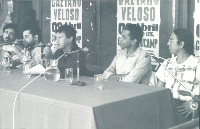 Foto em preto e branco de Tom Zé, José Carlos Capinam, Fernando Faro, Paulinho da Viola e Gereba Barreto, ao fundo cartazes dos evento.